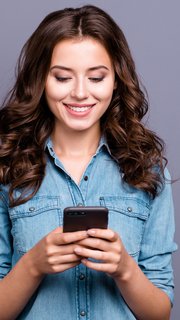 Content trendy cheerful nice cute adorable lovely attractive brunette girl with wavy hair in casual denim shirt, typing in phone, isolated over grey background