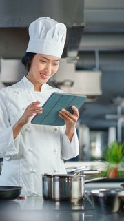 World Famous Restaurant: Asian Female Chef Cooking Delicious and Authentic Food, Uses Digital Tablet Computer While Working in a Modern Professional Kitchen. Preparing gourmet organic Dishes